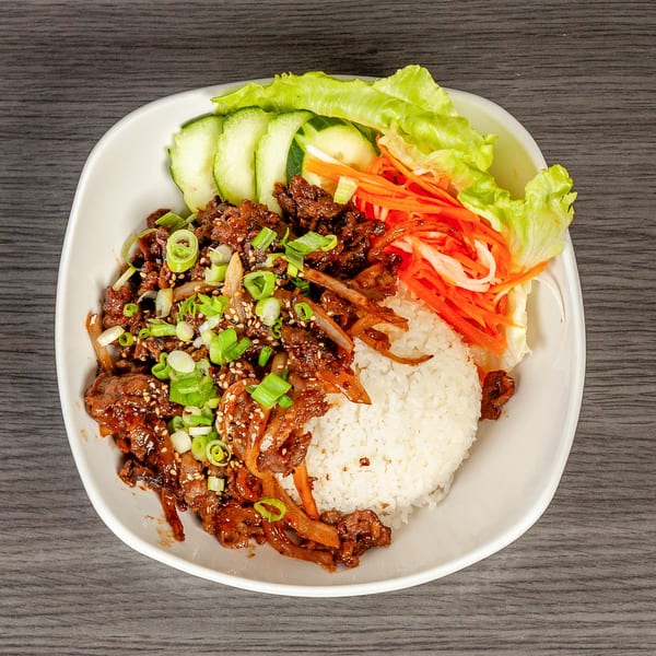 A white bowl filled with bulgogi, white rice, sliced cucumbers, lettuce, and shredded carrots.