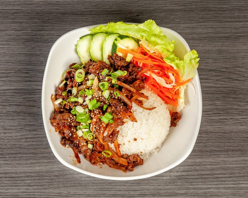 A white bowl filled with bulgogi, white rice, sliced cucumbers, lettuce, and shredded carrots.
