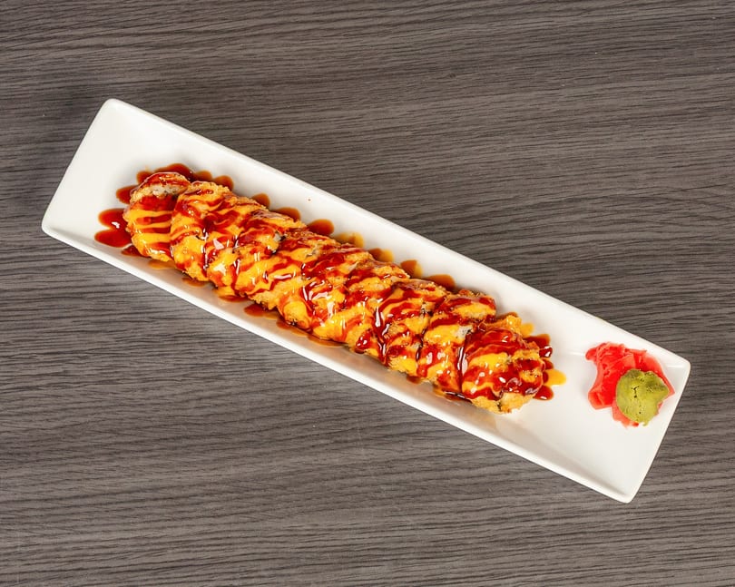Long white plate of deep-fried sushi rolls drizzled with orange and brown sauces, with ginger and wasabi.