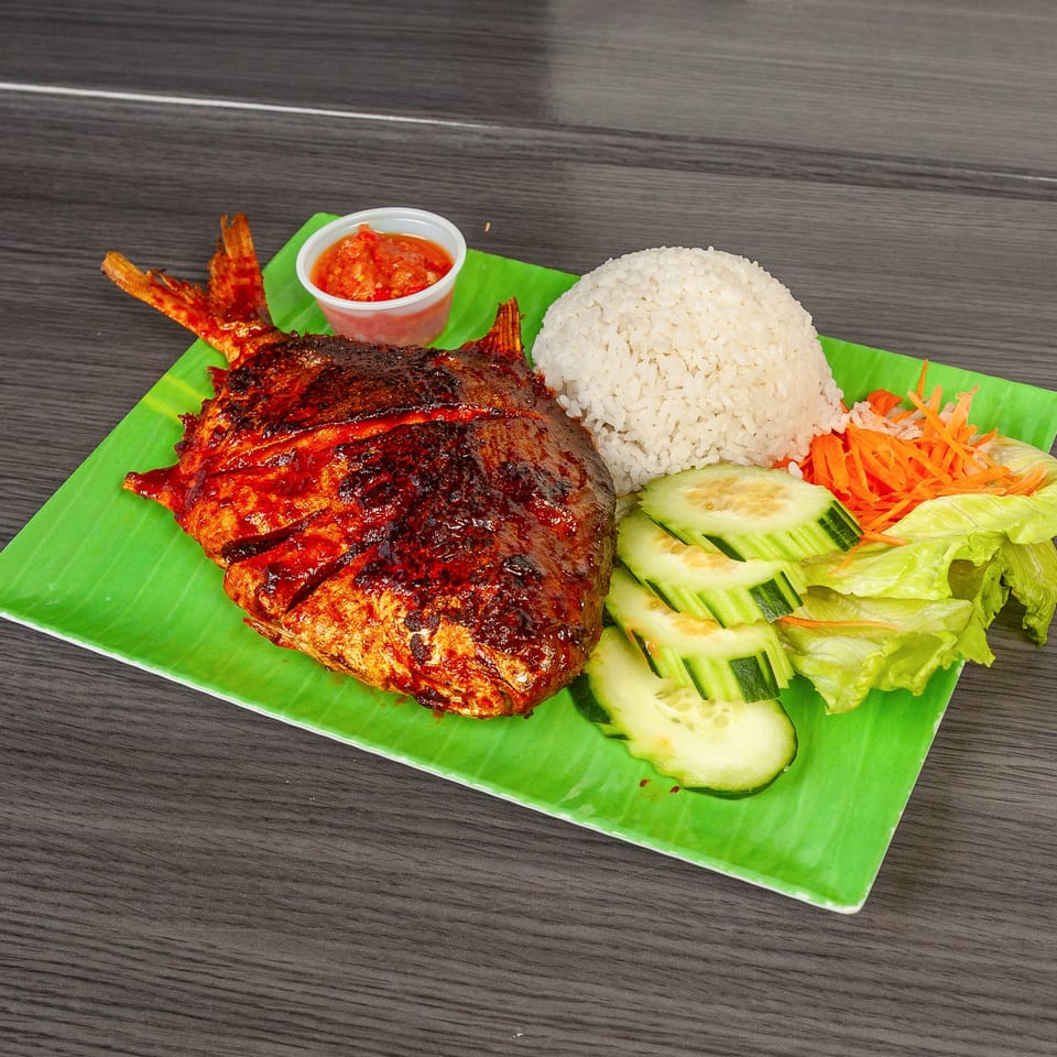 Grilled whole fish with rice, cucumber, lettuce, shredded carrots, and chili sauce on a green plate.