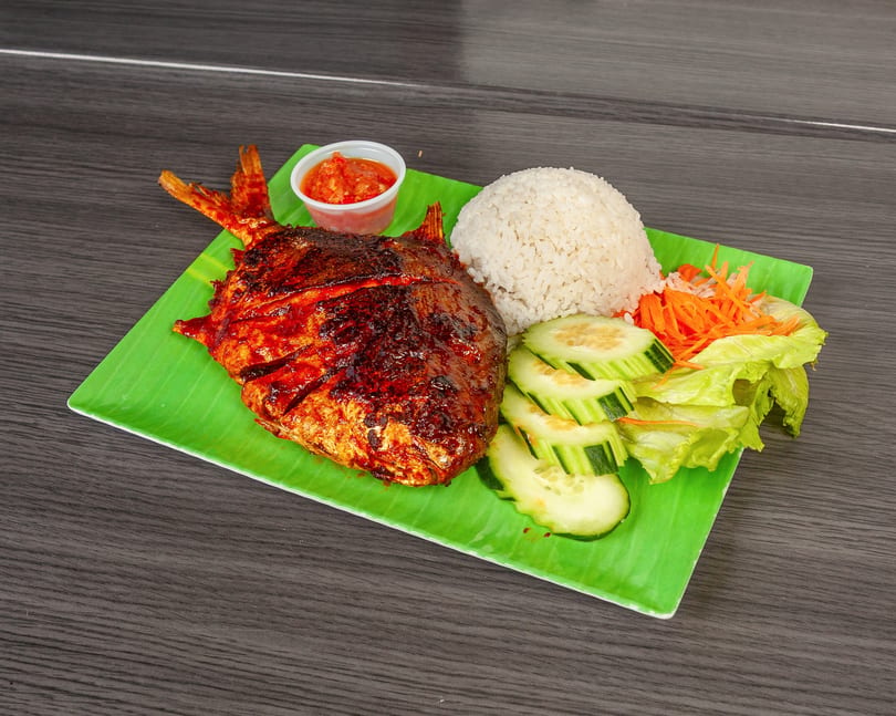 Grilled whole fish with rice, cucumber, lettuce, shredded carrots, and chili sauce on a green plate.