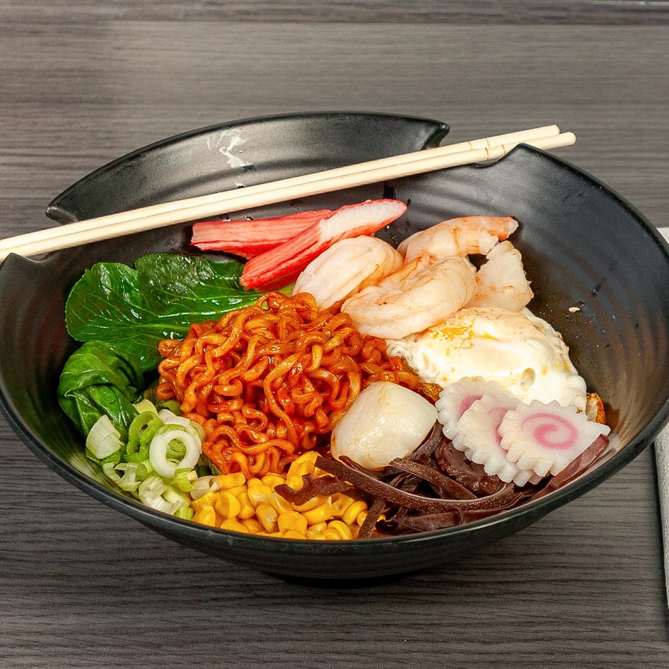 Spicy ramen bowl with shrimp, crab sticks, bok choy, corn, scallions, egg, and fish cakes.
