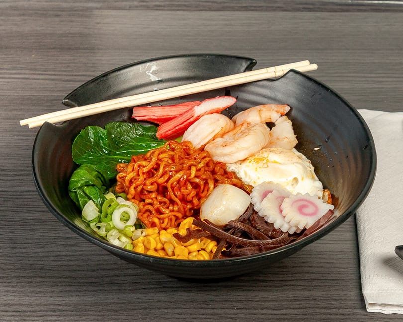 Spicy ramen bowl with shrimp, crab sticks, bok choy, corn, scallions, egg, and fish cakes.
