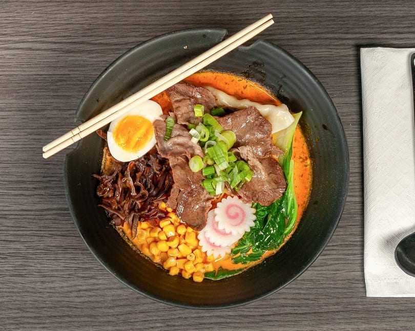 Overhead view of a ramen bowl with beef, egg, corn, scallions, and narutomaki.