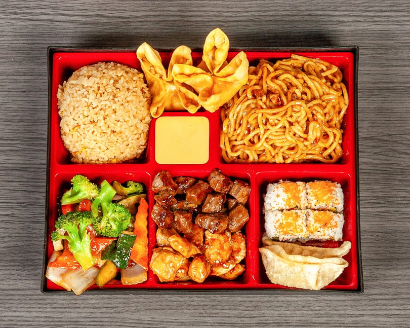 A red bento box filled with fried rice, lo mein, crab rangoon, vegetables, beef, chicken, sushi, and a dumpling.